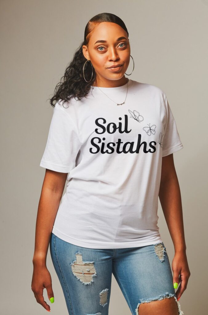 African-American woman wearing a white t-shirt with a delicate butterfly design and the term "Soil Sistahs" in script.